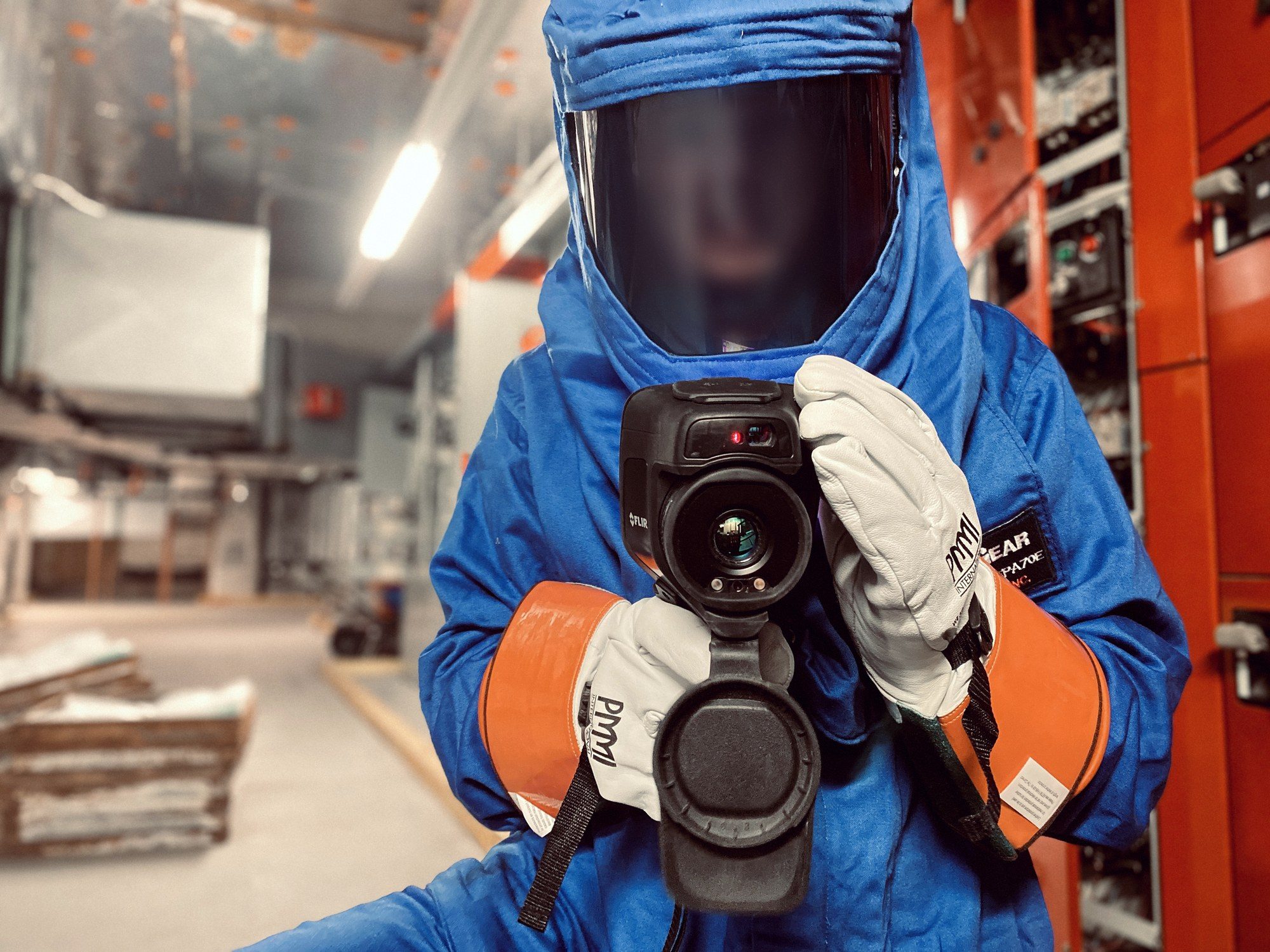 Thermographer in 40 cal/cm² arc-flash PPE aiming a FLIR thermal camera beside switchgear